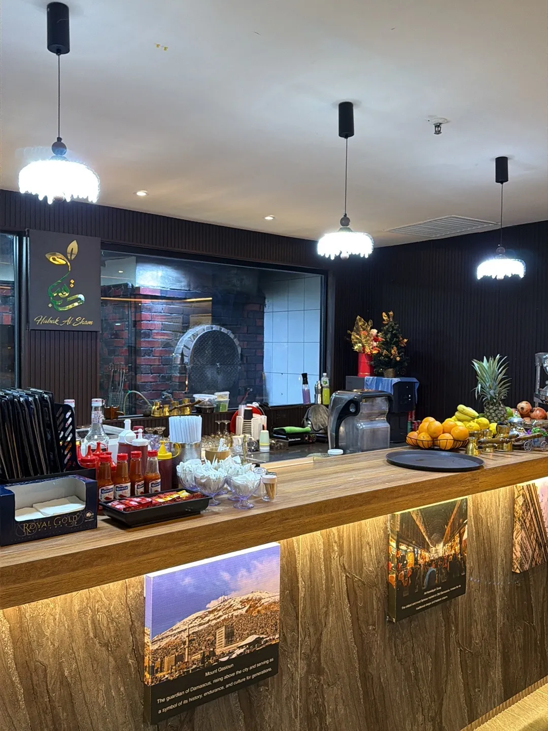 Counter with the Habak Al Sham logo, fresh fruit and Damascus prints, with the brick bread oven behind